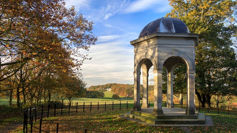 Small temple in the garden at Sheringham Park on a sunny day, surrounded by trees with hilly parkland in the background